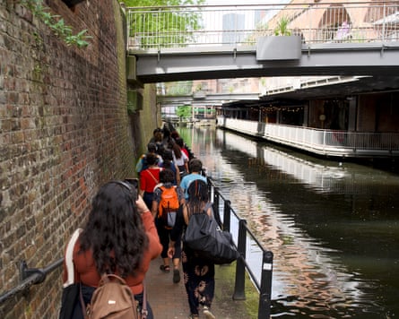 A view from behind the walking tour group as they follow a brick-lined canal path under a concrete pedestrian bridge.