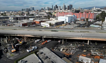 Overhead view of empty four-lane highway above industrial neighborhood with burned out vehicles visible below, under a blue sky.