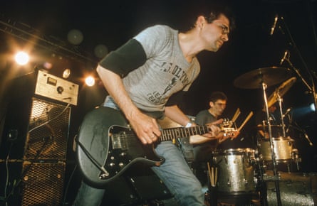 Steve Albini in a long-sleeved T-shirt and glasses playing guitar onstage with Shellac, the drummer behind him