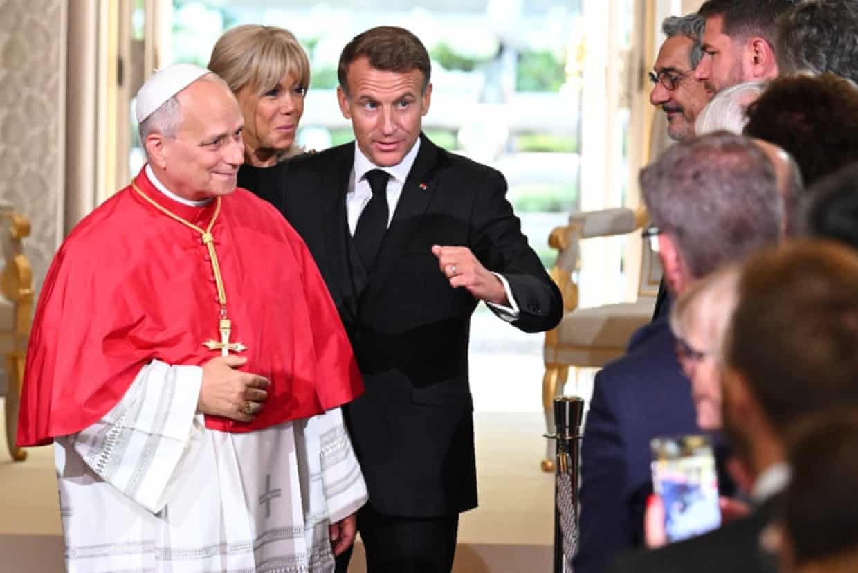 Macron walks the pope past a line of people inside building with exterior visible behind through doorway