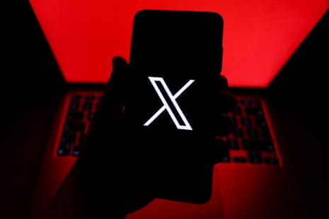 A silhouette of a hand holds a black phone with a white X social media platform logo in front of a red laptop screen
