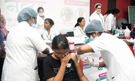 A girl sits on a chair and covers her mouth while a healthcare worker is poised to vaccinate her. The are surrounded by other healthcare workers at what seems to be a mass vaccination event.