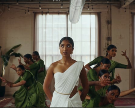Women wearing green dresses dancing behind a woman in a white dress in a sparsely decorated room