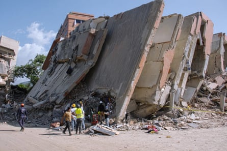 A small group of rescue workers stand around a collapsed building, where the walls have tumbled against one another and slanted like falling dominoes.