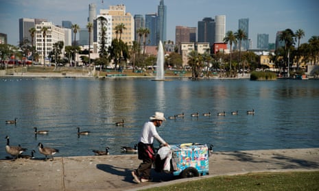 A man pushes an ice-cream cart next to a lake with the downtown Los Angeles skyline in the background.