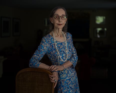 Joyce Carol Oates in glasses, pearls and a blue patterned dress stands beside a cane chair in a dark room