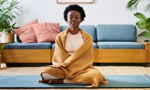A young Black woman sitting on a yoga mat in her lounge at home meditating.