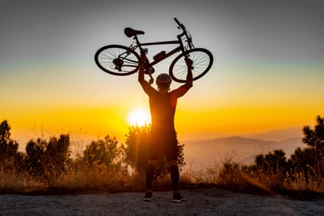 Male biker holds bike above head after successfully biking up a hill at sunset
