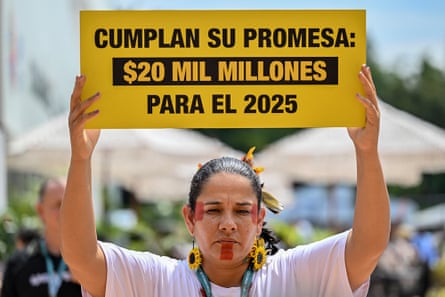 An Indigenous woman wearing red face paint holds a sign in Spanish above her head.