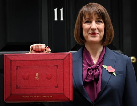 Rachel Reeves, in a bow-tie shirt and jacket with a poppy on the lapel, holds up a budget wooden box with a handle, smiling