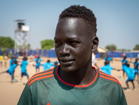 Portrait of Alaak ‘Kuku’ Akuei outside, with a group of young people holding hands in a circle behind him