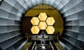 Man in a spacesuit-like jumpsuit stands, hands on hips, in from of hexagonal shapes in a metal tunnel that form part of the James Webb space telescope