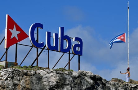 A tourist takes a selfie picture as she stands next to a Cuban national flag and a sign comprised of the letters spelling out Cuba