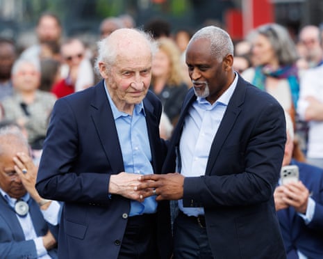 Pierre Guillon de Princé, an elderly white man, stands with Dieudonné Boutrin, a middle-aged black man with grey hair, at an event to unveil a memorial. They both wear dark blue suits and open-necked pale blue shirts and hold hands to each other's backs and touch hands in front.