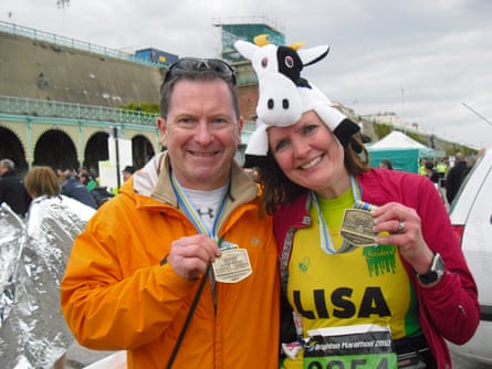 A man in an orange anorak and a woman with a yellow T-shirt with her name LISA on it and a black and white cow hat on her head - both holding up medals standing close together from waist up