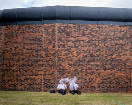 Two people sitting on grass and vaping, as they lean against a large brick wall.