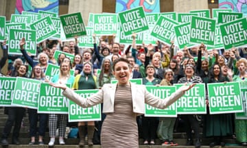 Carla Denyer smiles with her arms outstretched. A large group of people stand behind her holding large signs that bear the words 'Vote Green'.