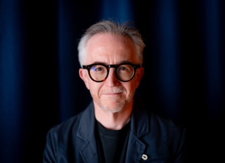 Man with grey hair and black-rimmed glasses wearing dark blue blazer against a similarly coloured backdrop