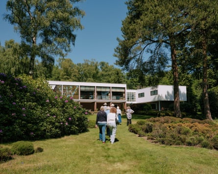 A flat-roofed two-storey white house with many floor-to-ceiling windows in a garden with a lawn, trees and shrubs