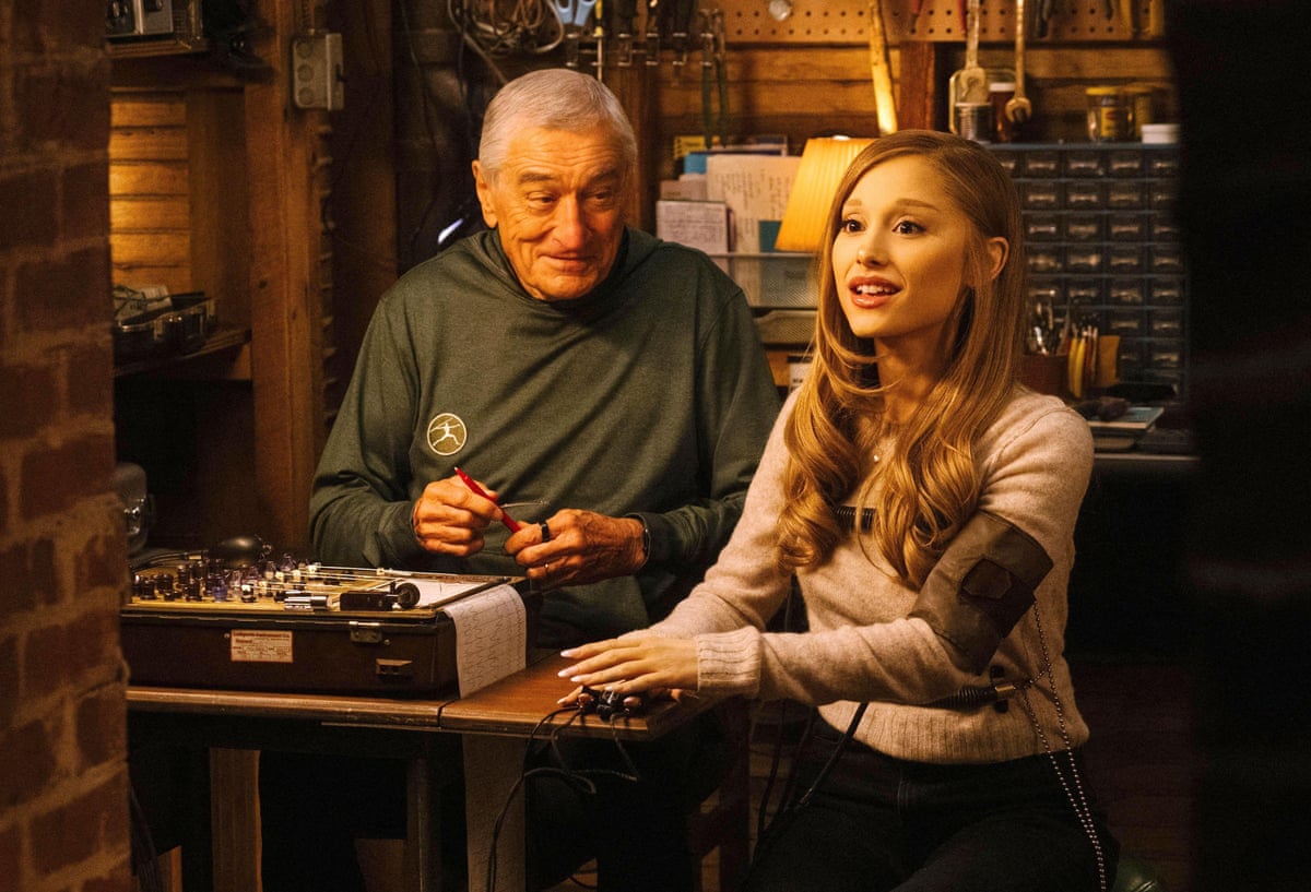 Ariana Grande sits beside Robert De Niro with a cuff around her upper arm connected to a machine spewing out a graph of some kind.