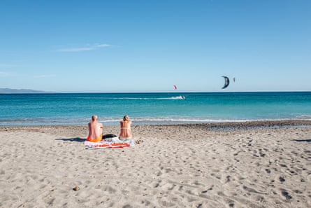 a couple stting on a white sand beach with blue sea and sky, windsurfers on the horizon