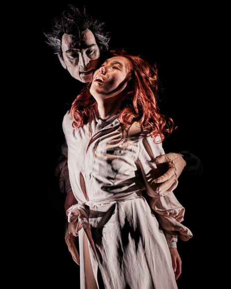 A puppet with wild red hair and white masks on their chest raises their arms while surrounded by actors in period costume in Dracula: Lucy’s Dream.