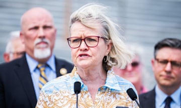Middle-aged white woman, blond hair, red glasses, collared yellow and blue buttoned blouse, long pearls, speaks into microphone in front of group of white men as wind sweeps hair.