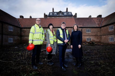 Two men and two women stand in courtyard with three walls of the historic brick building around them; two are carrying red safety helmets and three are wearing hi-vis vests or jacket.