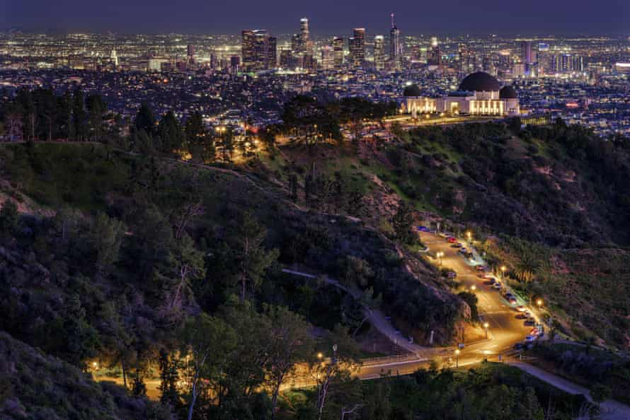 Griffith Park is seen at dusk in Los Angeles. The main road through the park and domed Griffith observatory building glow with warm light agains a background of the Los Angeles skyline.