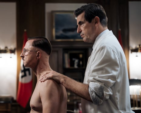 A shirtless Martin Müller as Heinrich Himmler sits while Claes Bang as Felix Kersten in a white shirt examines his shoulder from behind