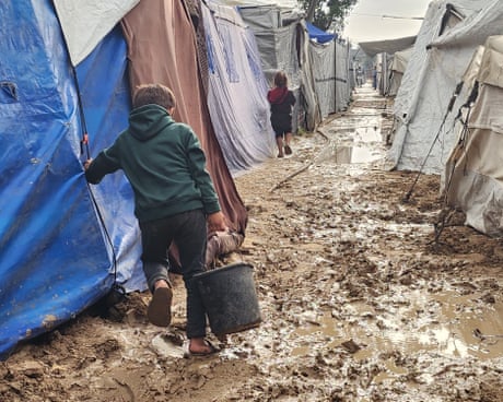 A boy seen from behind walks through mud between tents.