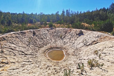 A huge crater with a small amount of brown water at its centre, with some plants growing in the rocky, ridged surrounds.