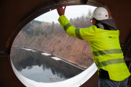 A man in a high-vis jacket looks out of a round window at a lake