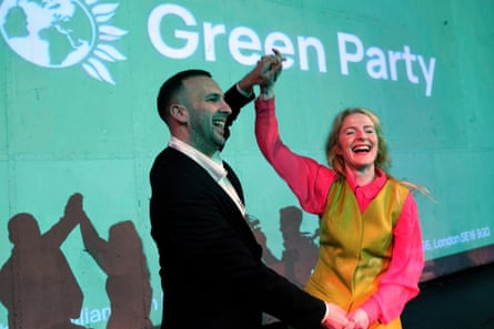 They smile broadly while holding hands and gesticulate towards an off-camera crowd, standing in front of a screen that reads ‘Green Party’