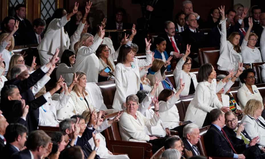 Female members of Congress wearing white, hold up three fingers for the HR3 health care bill as Donald Trump talks about healthcare during his State of the Union address.