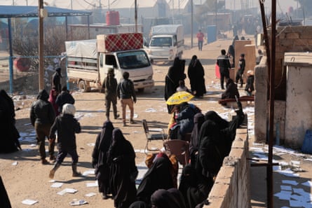 Groups of people including women and children sit and stand around a wall and on a track between shelters; there are heavily laden small trucks on the path. The scene appears quite chaotic.
