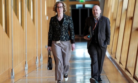 Lorna Slater and Patrick Harvie in a corridor at Holyrood; she is tall and has curly fair hair, and wears loose silky trousers with a black jacket that has a gold leaf-like pattern; he is shorter and wears a dark grey suit plus a rainbow-striped lanyard.