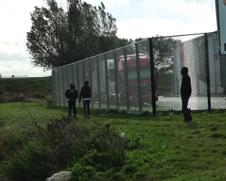 Migrants stand near a wire fence beside parked lorries at a depot in Calais