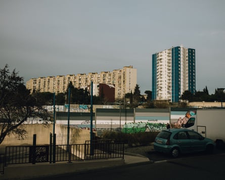 In Marseille’s Malpassé neighbourhood, a car and van sit near a graffiti wall, with housing towers beyond