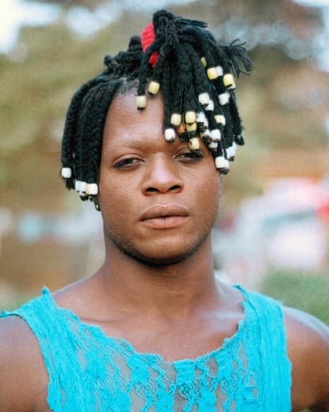 A person in a turquoise lace vest looks directly at the camera, with braided hair decorated with white and yellow beadshave stopped time” A Family Portrait