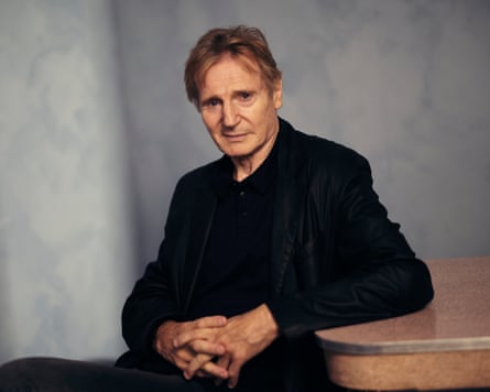 Liam Neeson sits at table in dark clothes and faces the camera