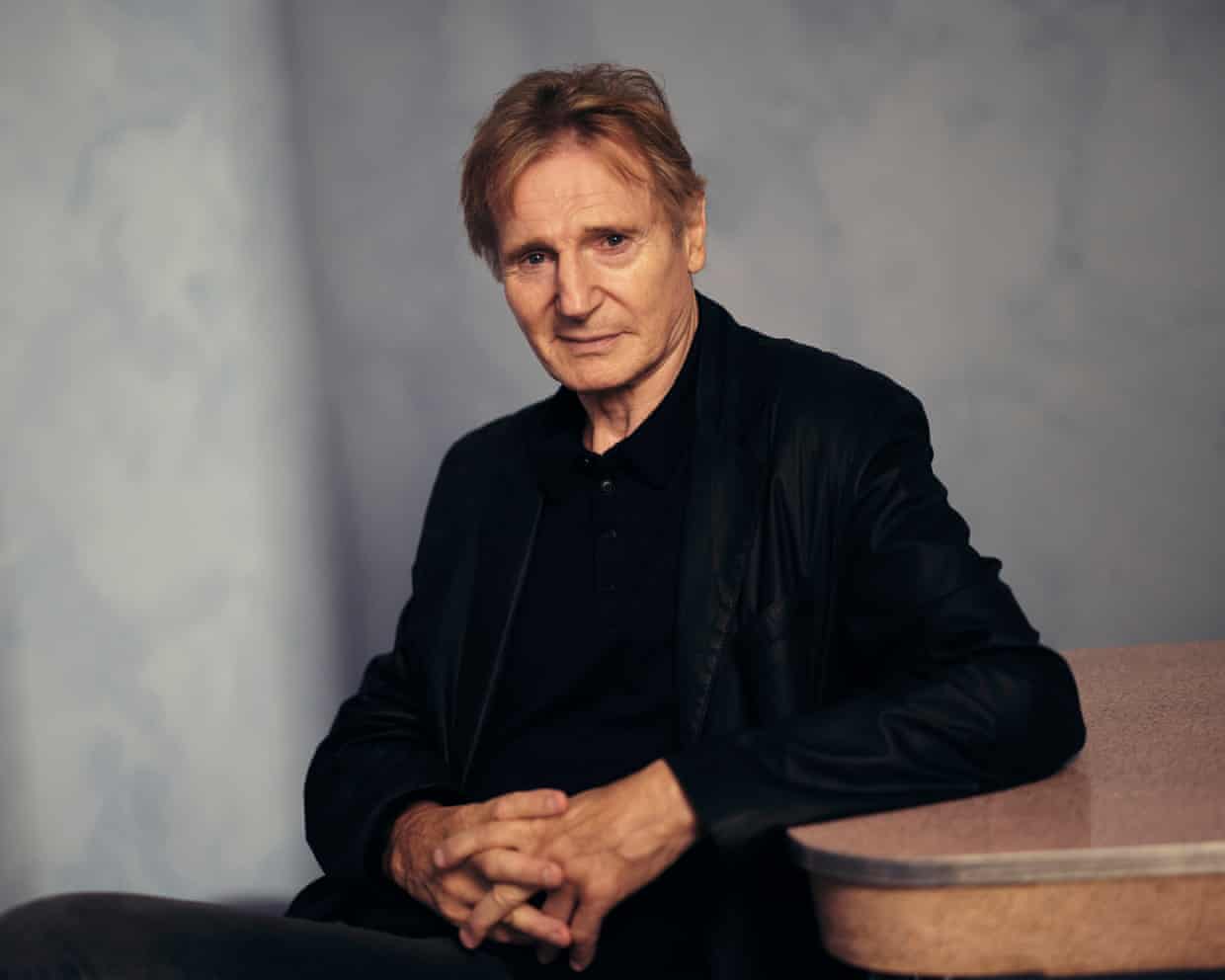 Liam Neeson sits at table in dark clothes and faces the camera