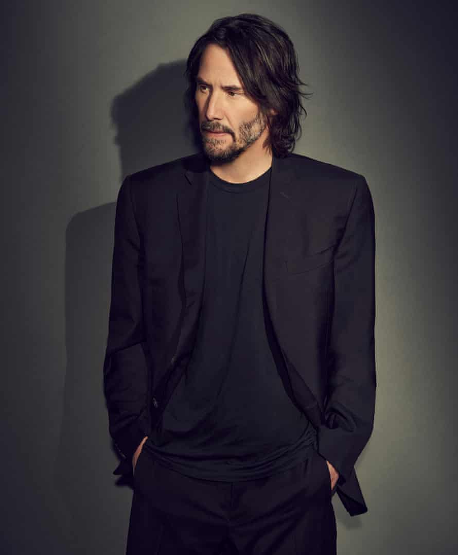 Keanu Reeves in a black suit and black t-shirt, looking to his right, hands in pockets