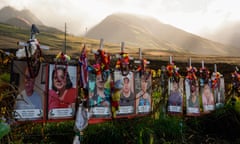 A row of color photographs of people, with ribbons and a white cross attached to each, on a fence, with the sun setting behind distant hills.