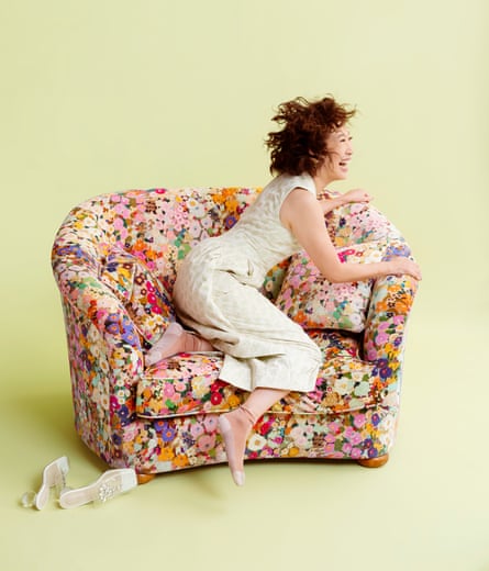 Oh wears a white textured dress while laughing and sat on a floral sofa with her heeled shoes sitting by her side