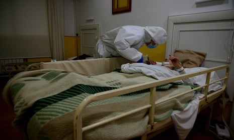 A doctor wearing a face mask and protective clothing attends to an elderly woman lying in a bed with safety rails