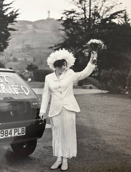Woman in cream suit with feather hat raises bunch of flowers next to car that reads ‘just married’
