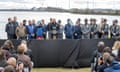 man speaks at podium amid officers outdoors, with the water in the background