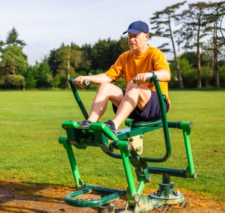 A man in shorts and a cap sat on an outdoor rowing machine in the park