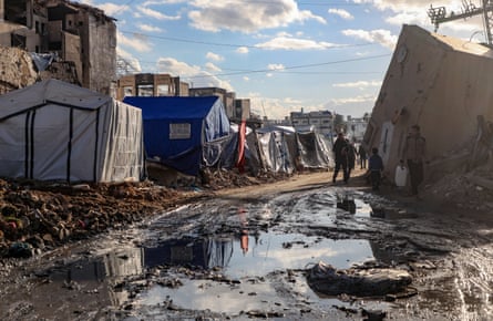 People walk past tents erected alongside ruined buildings, as large puddles sit on the road